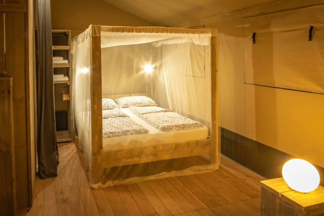 Schlafzimmer der Wood Lodge für 2 bis 6 Personen mit Himmelbett unter einem Moskitonetz, offenem Schrank mit Handtüchern und einer Lampe auf dem Nachttisch