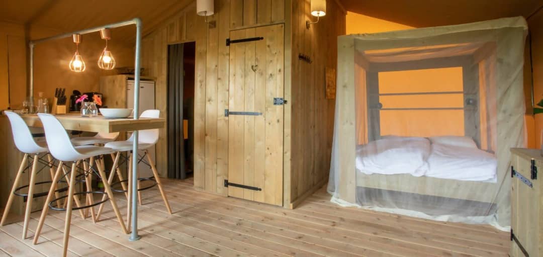 Wohnbereich der Desert Lodge XL mit Holzbar, weißen Barhockern, Holztür und Himmelbett mit Moskitonetz