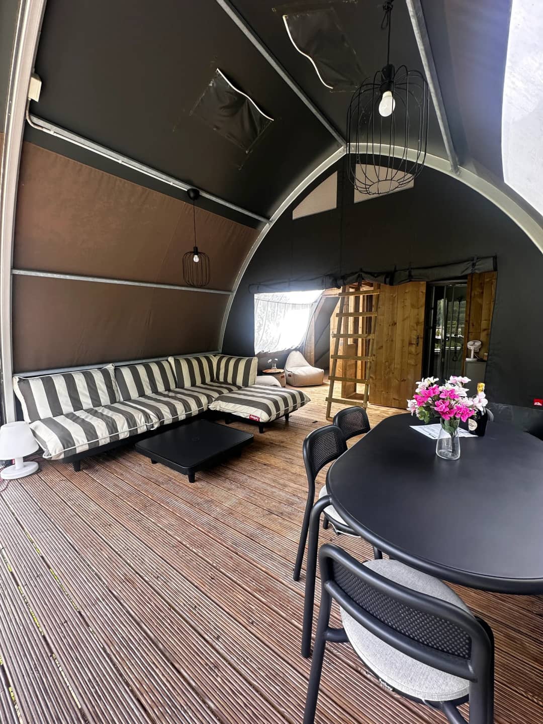 Wohnbereich der Big Oak Lodge XL mit gestreiftem Loungesofa, schwarzem Esstisch mit Blumen und einer Holzleiter