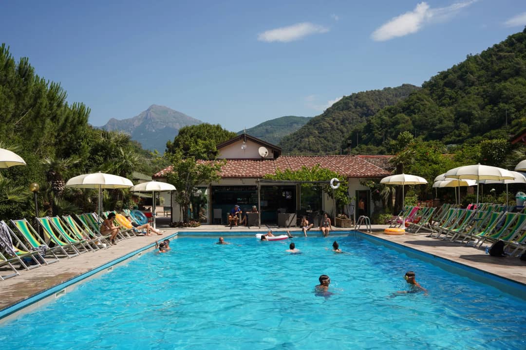 Der Pool vom Campingplatz Delle Rose mit grün-weißen Liegestühlen, weißen Sonnenschirmen und einem Berggipfel im Hintergrund