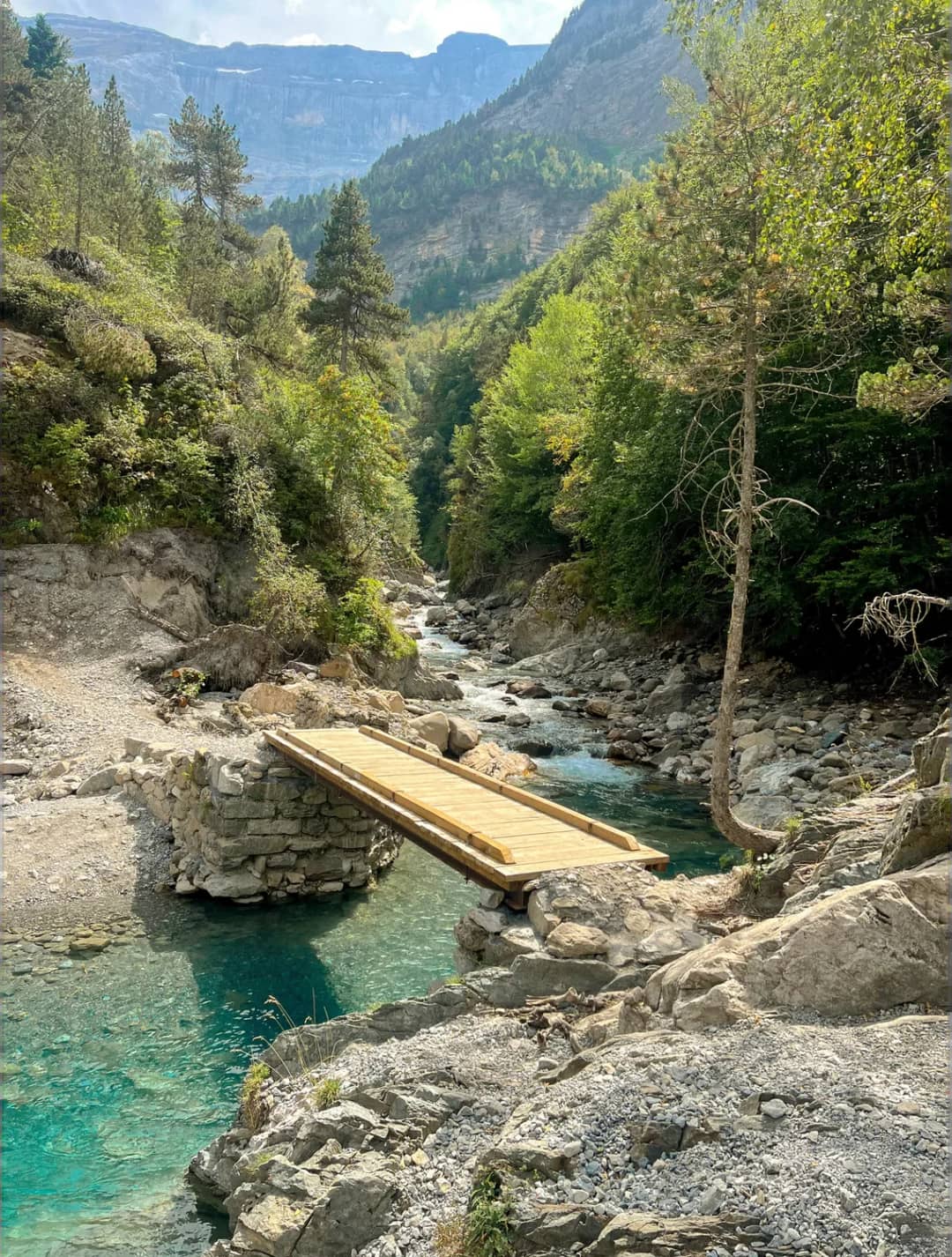 Klarer blauer Gebirgsfluss mit Holzplattform in einem waldreichen Tal, umgeben von Felsen und Nadelbäumen, Berge im Hintergrund.